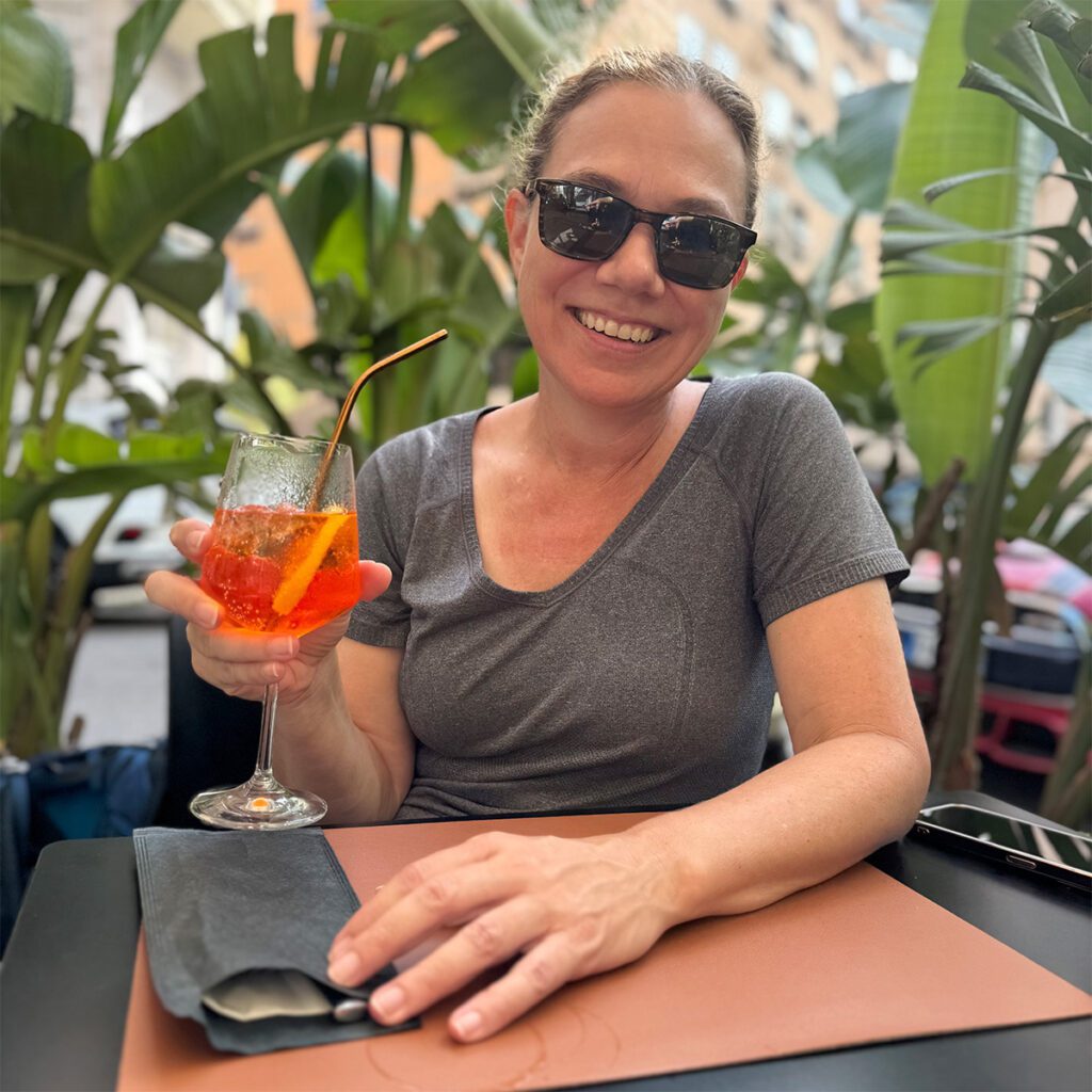 Yvette enjoying an Aperol Spritz in Rome, holding the vibrant orange cocktail with a warm smile.